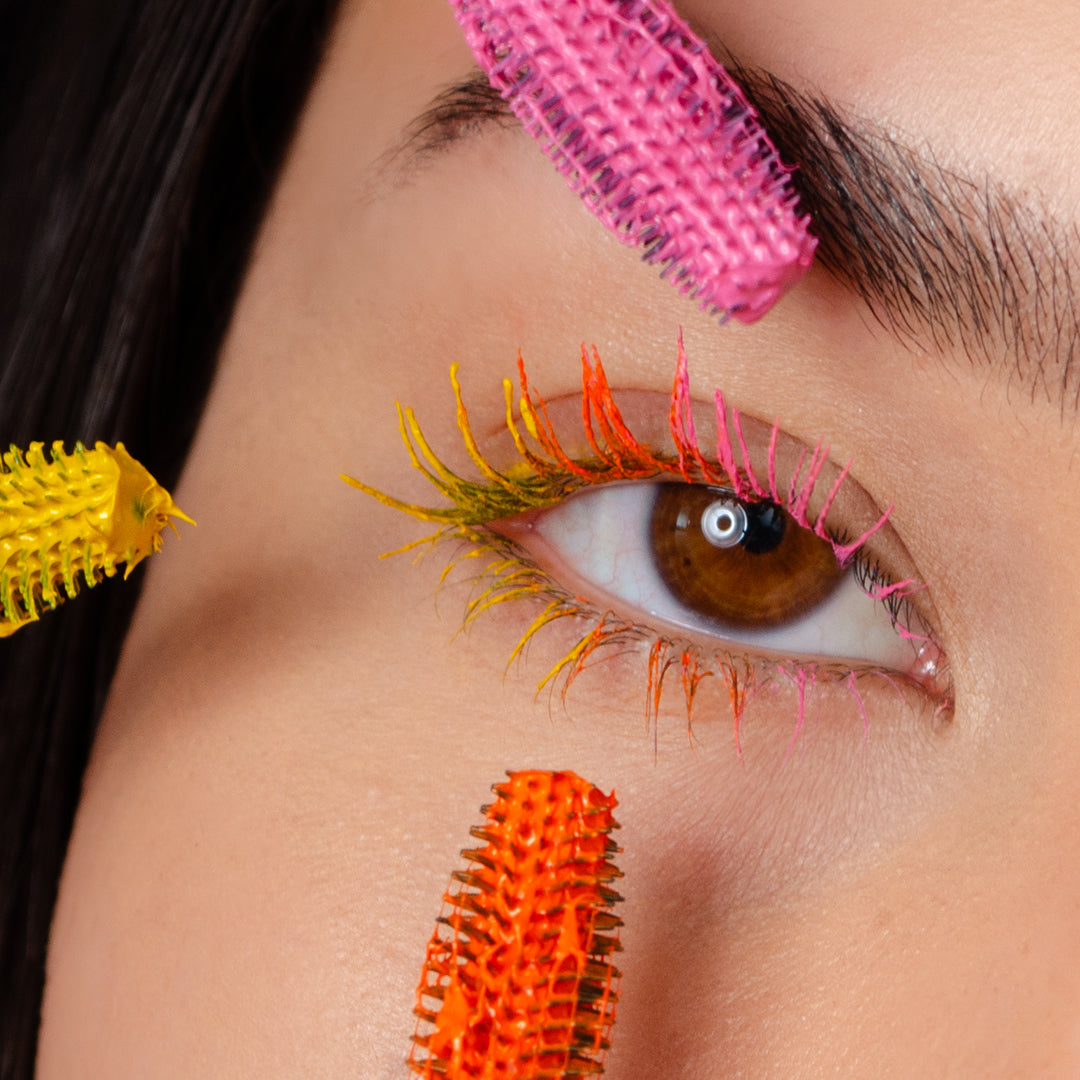 A close-up of a brown eye with multi-colored mascara on the lashes and several colorful mascara wands near the eye, highlighting vibrant makeup.