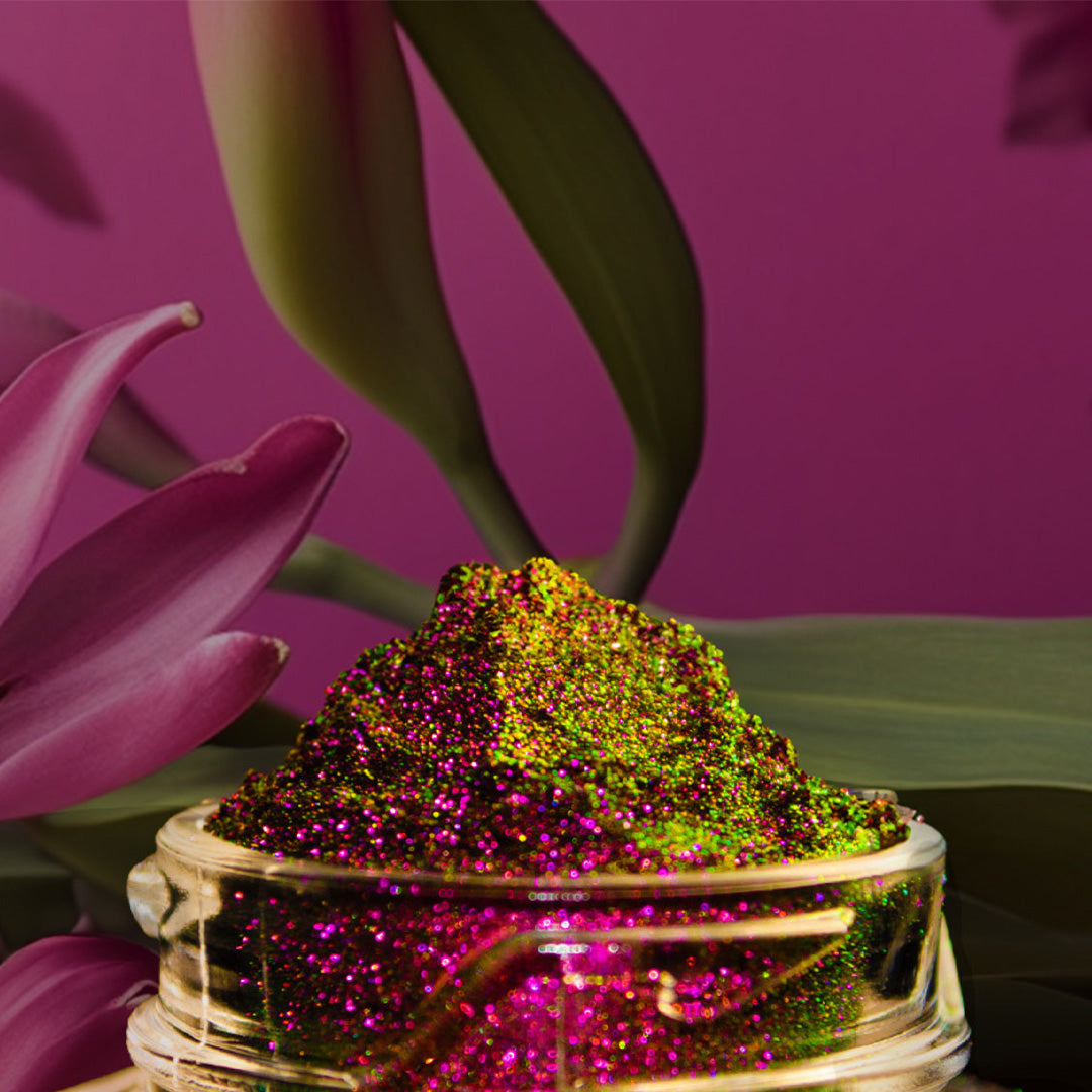 A small jar filled with vibrant, sparkling pink and green glitter sits beside pink and green flower petals against a pink background.