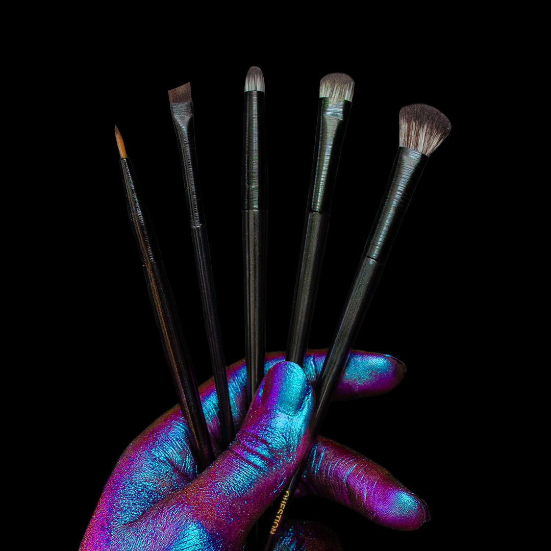 A hand covered in shiny, colorful paint holds five makeup brushes fanned out against a solid black background.
