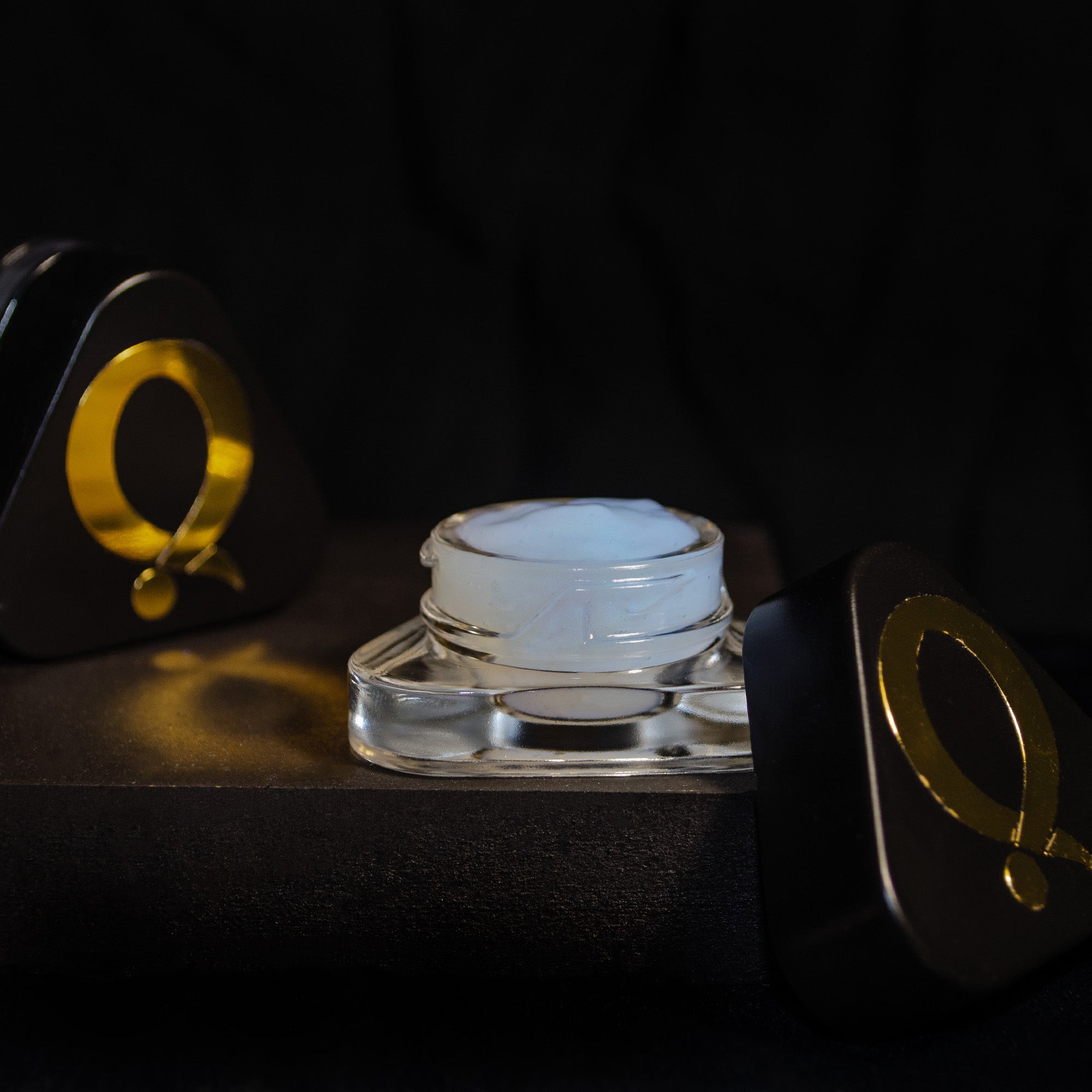 A small glass jar of white cream sits on a dark surface, flanked by two black and gold containers with a circular design, set against a black background.