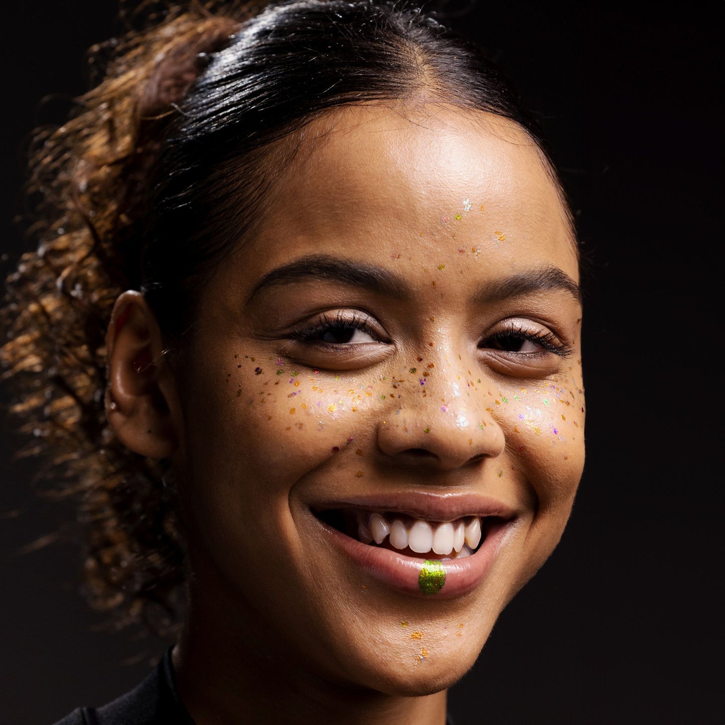 A woman with curly hair smiles, showing glitter on her face and lips in front of a dark background.
