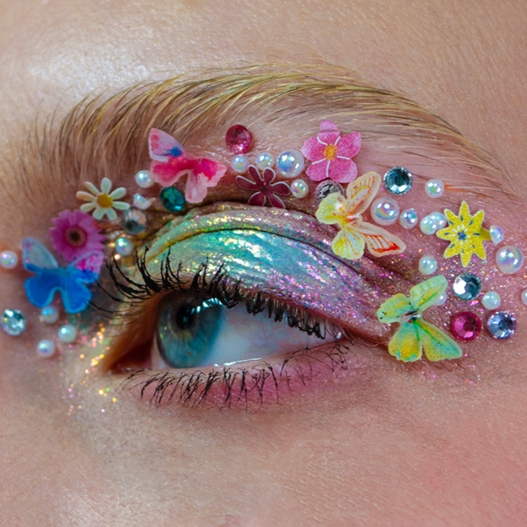 Close-up of a blue eye with colorful glitter eyeshadow, rhinestones, and butterfly decorations above the eyelid.
