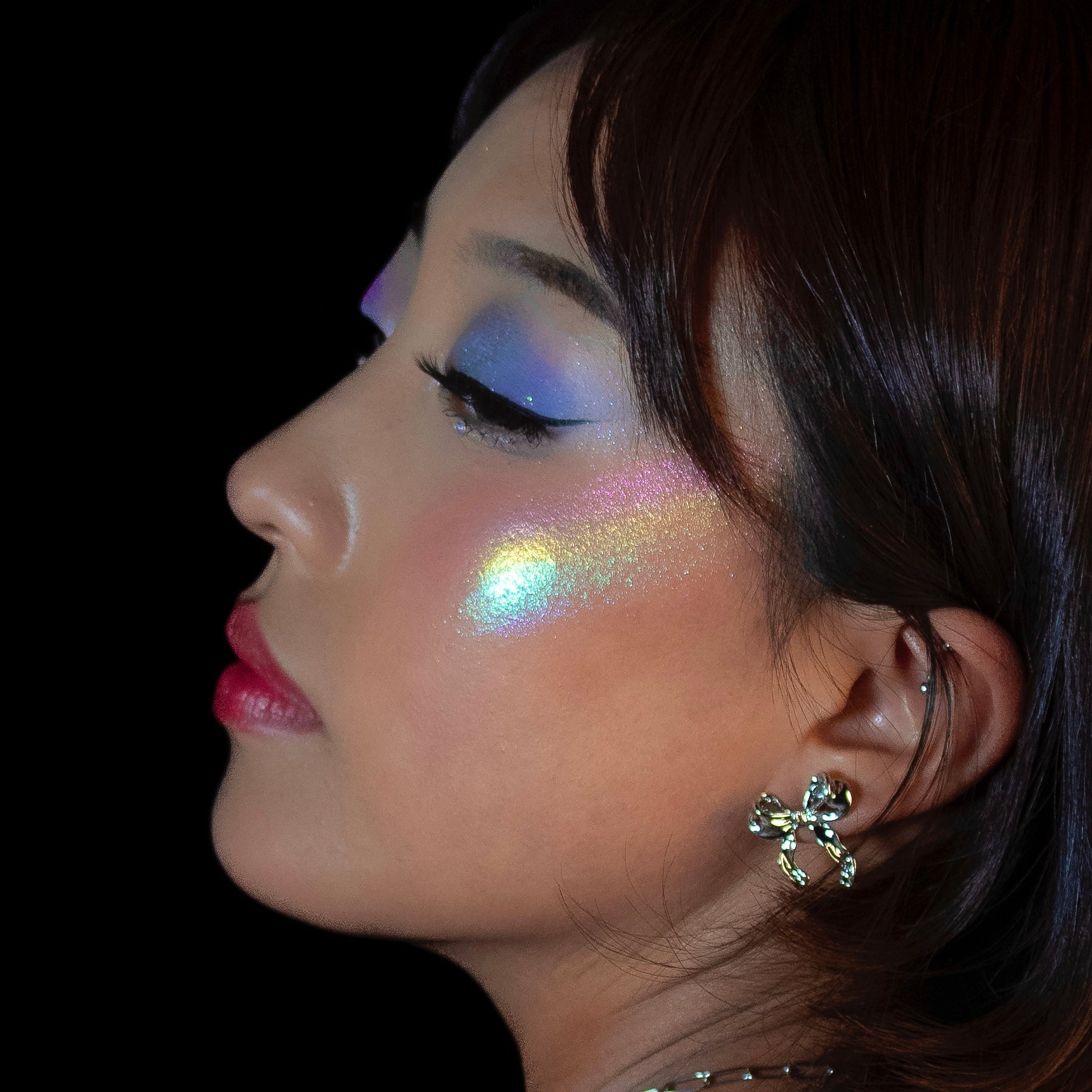 Woman in profile with highlighted cheekbones, winged eyeliner, red lips, and a silver bow earring on a black background.