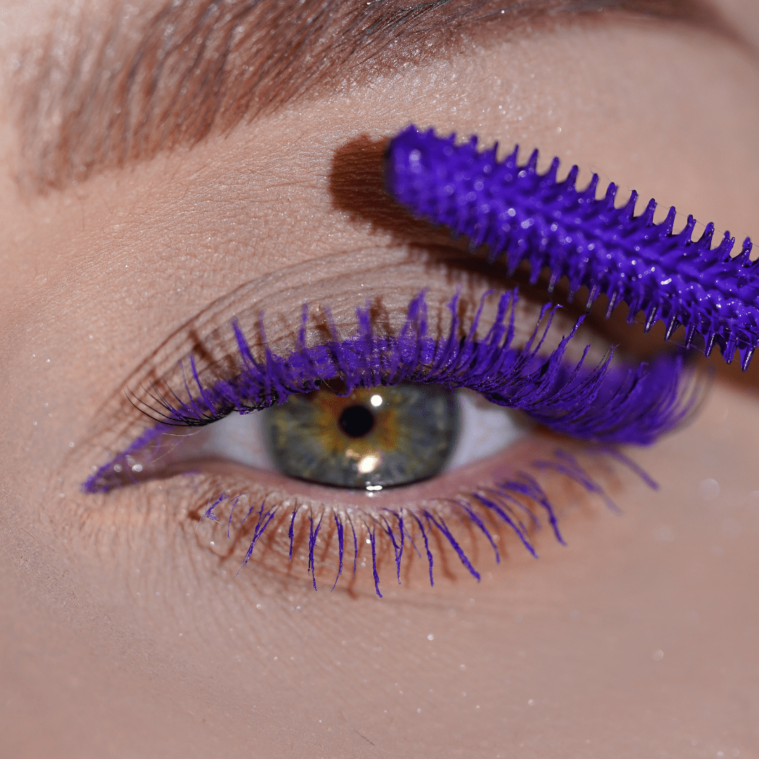 Close-up of a green eye with purple mascara applied to the upper lashes using a vibrant purple wand