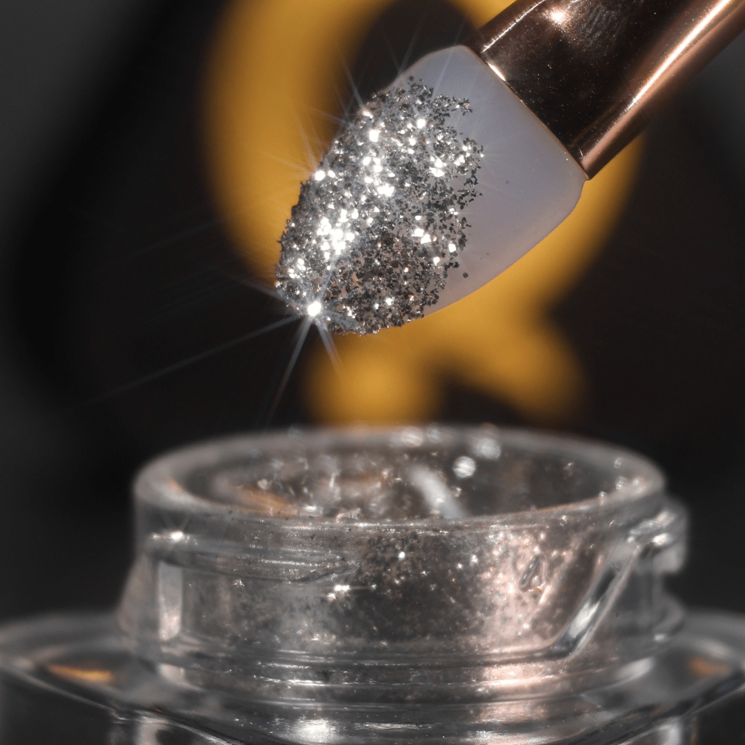 A brush holds silver glitter powder above an open jar, with the powder visibly sparkling in the light.