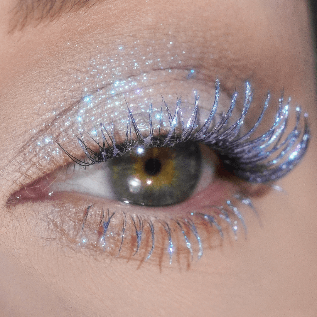 Close-up of an eye with silvery blue glitter on the eyelid and visible Mascara Topper - Frosty Lilac on the lashes.