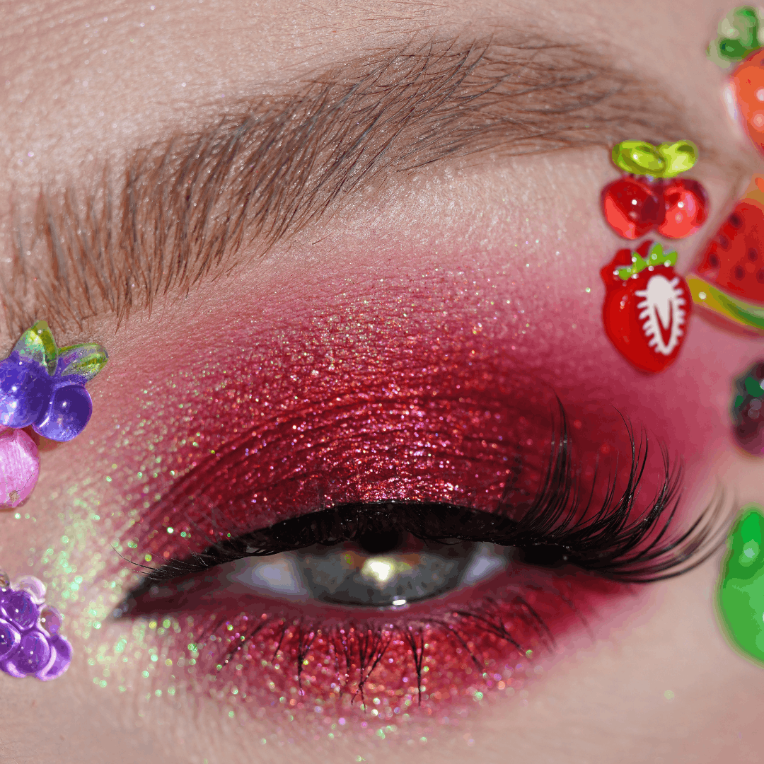 Close-up of an eye with red glitter eyeshadow, long lashes, and small colorful Fruit Gems near the eyelid.