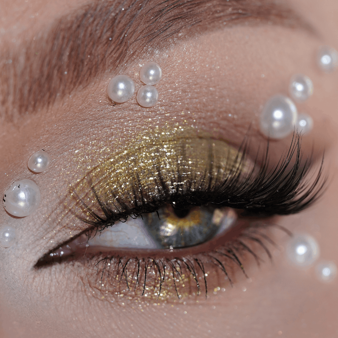 Close-up of an eye with gold glitter eyeshadow, dramatic lashes, and self-adhesive white pearls on the eyelid.