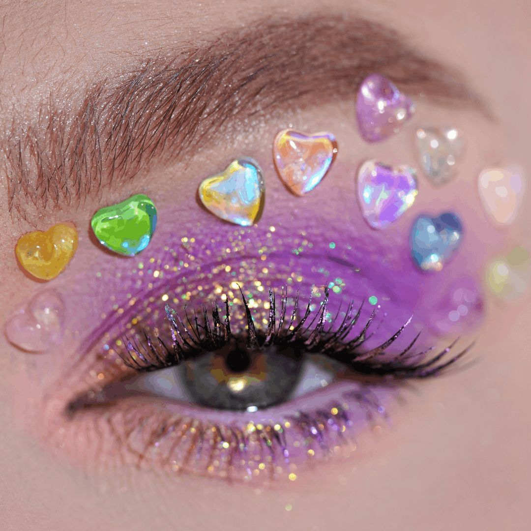 Close-up of an eye with purple and gold glitter eyeshadow, long lashes, and crystal heart-shaped gems above the eyelid.