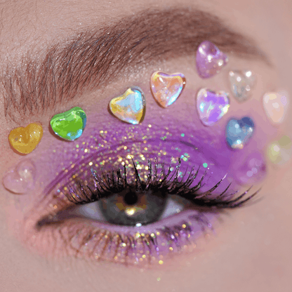 Close-up of an eye with purple and gold glitter eyeshadow, long lashes, and crystal heart-shaped gems above the eyelid.
