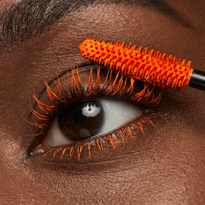 Close-up of an eye with orange mascara being applied to the upper lashes.