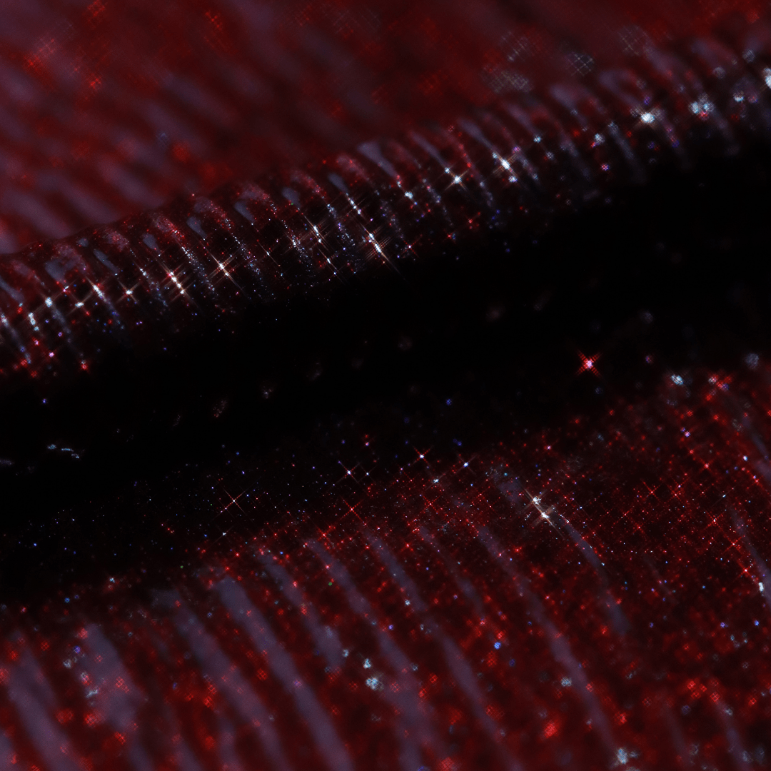 Close-up of a mascara applicator coated in Dark Flame showing shimmering red color with glitter highlights and a ribbed texture.