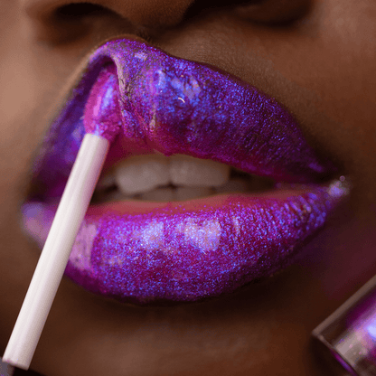 Close-up of lips with glossy purple and blue glitter, a gloss applicator touching the lips.