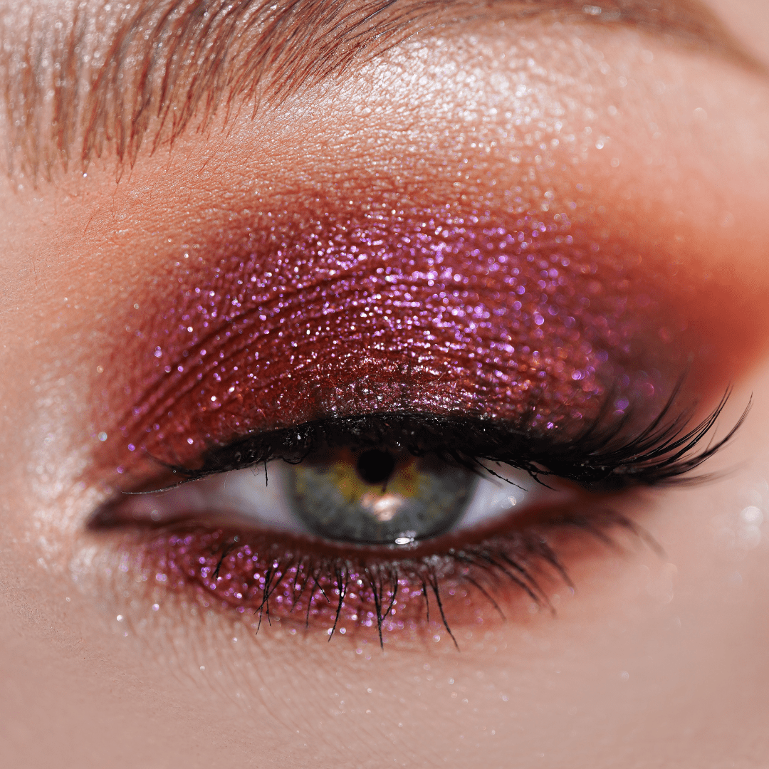 Close-up of an eye with sparkly pink and dark red eyeshadow, winged eyeliner, shimmer accents, and thick lashes.