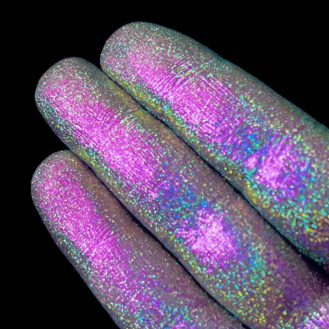 Three fingers hold shimmery pink and blue loose pigment against a black background.