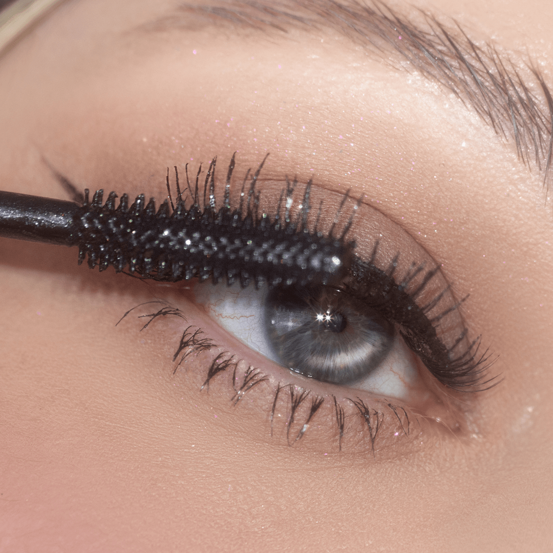A black mascara wand applies mascara to upper lashes near a blue eye in a close-up shot.