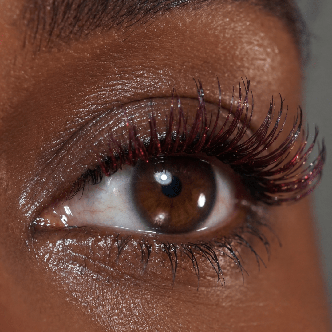 Close-up of a brown eye with long lashes coated in dark mascara with red shimmer on the tips.