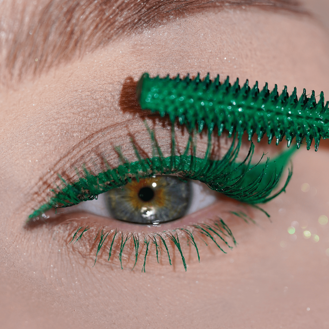 A close-up of an eye shows lashes being coated with green mascara using a green wand.