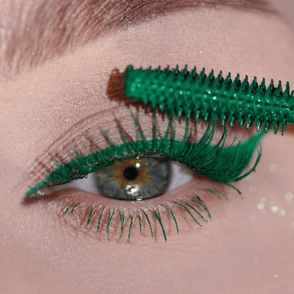 A close-up of an eye shows lashes being coated with green mascara using a green wand.