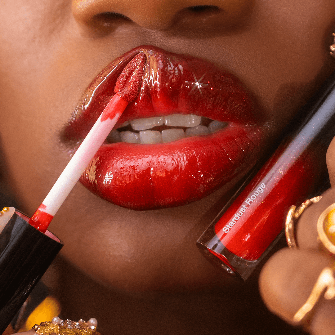 A hand holds a visible tube labeled Stardust Rouge while applying lip gloss.