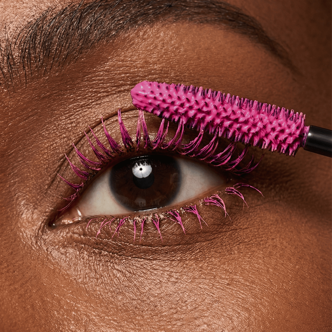 A person applies bright pink mascara to their upper lashes using a matching pink wand labeled Lotus Bloom.