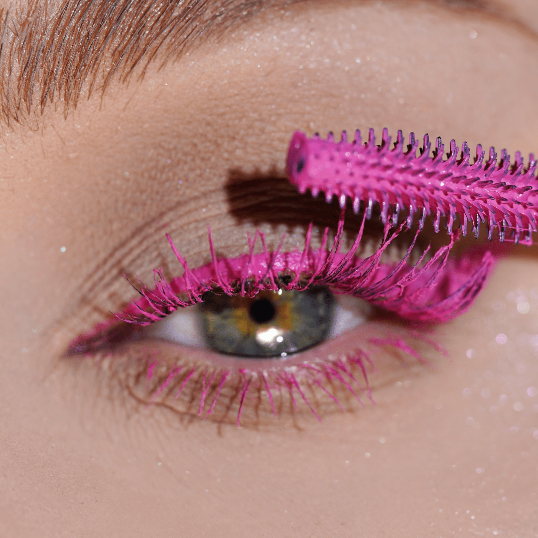 Close-up of an eye with a wand applying pink mascara to the upper lashes