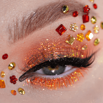 A close-up of an eye with orange glitter eyeshadow, red and orange gem accents, and bold black lashes.
