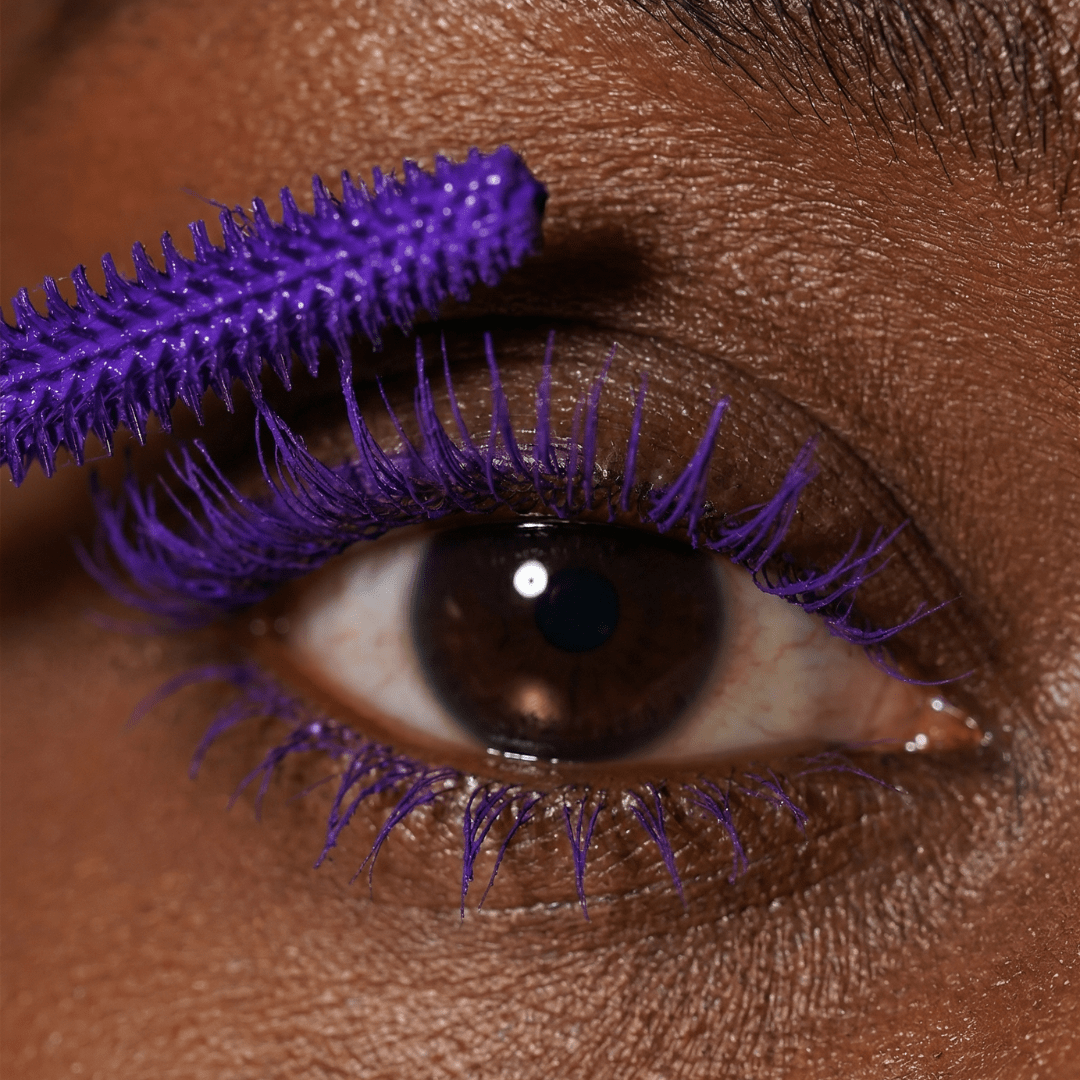 Close-up of a brown eye with purple mascara on the upper lashes and a matching purple wand applying it.