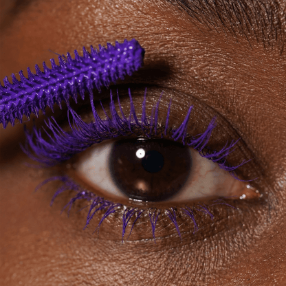 Close-up of a brown eye with purple mascara on the upper lashes and a matching purple wand applying it.