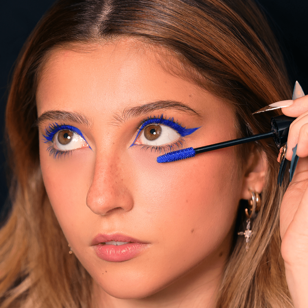 A woman applies blue mascara to her lower lashes with matching blue eyeliner and bold color on her upper eyelids.