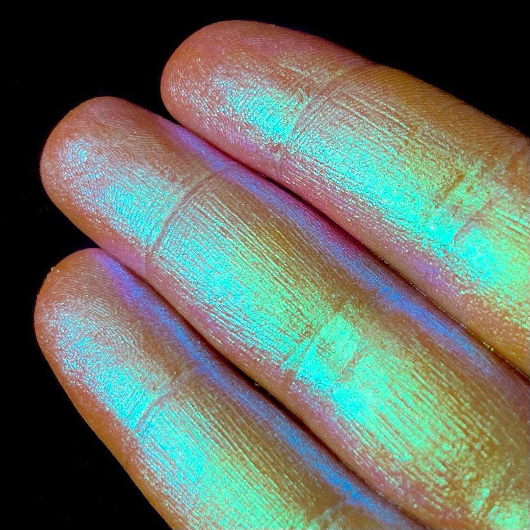 Three fingers display iridescent gold-pink pigment on their nails against a solid black background.