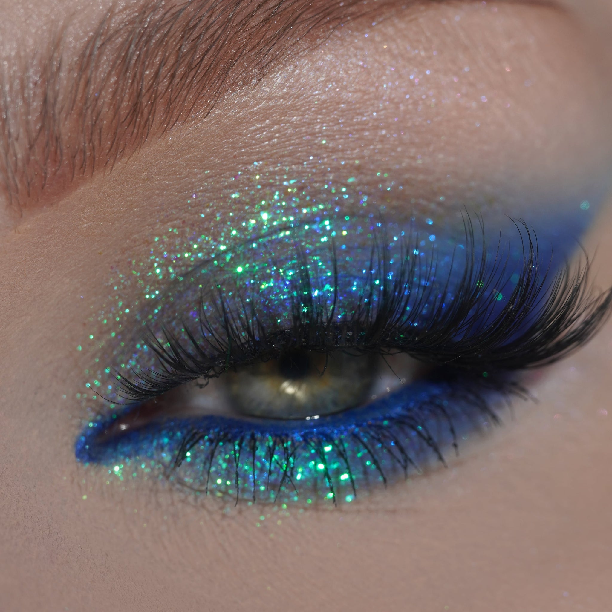 Close-up of an eye with blue and green glitter on the eyelid, and long black eyelashes.