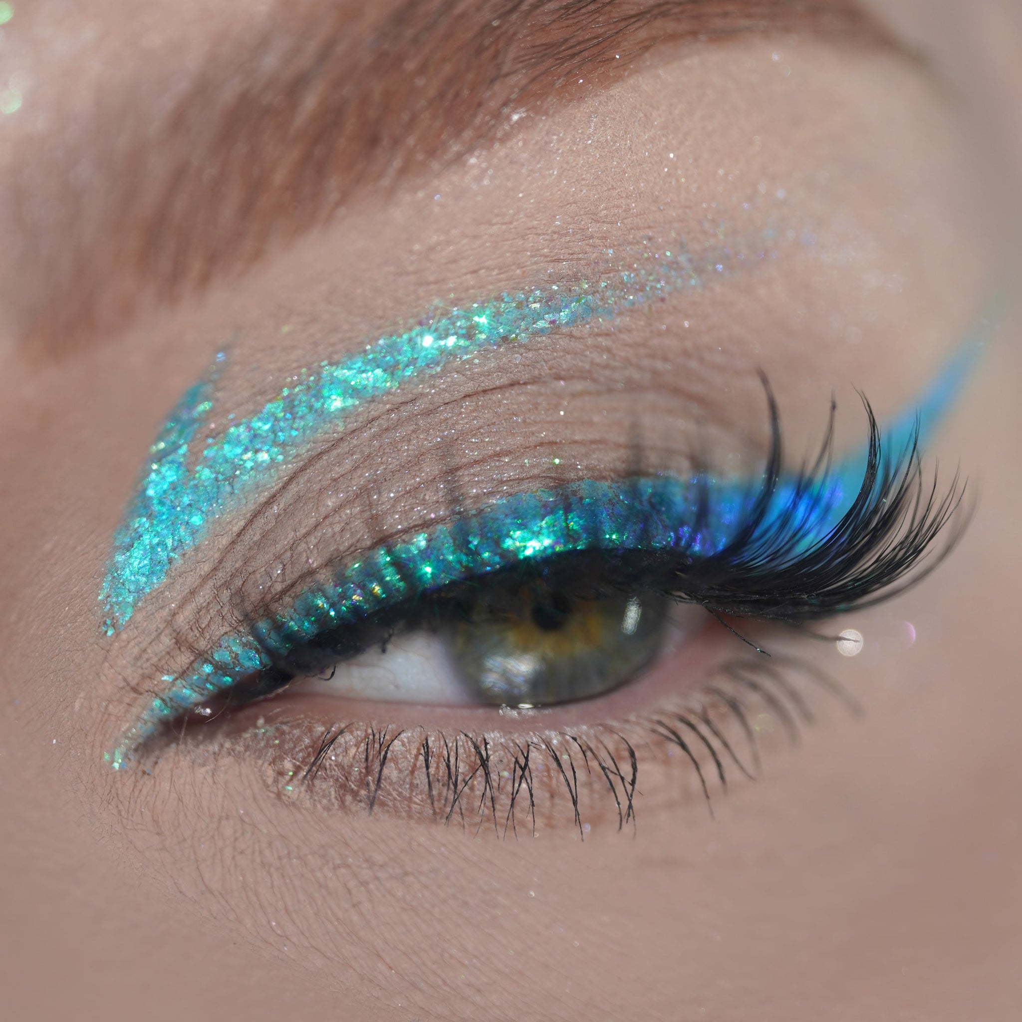 Close-up of an eye with glittery turquoise liner on the eyelid, long eyelashes, and a natural brown eyebrow.