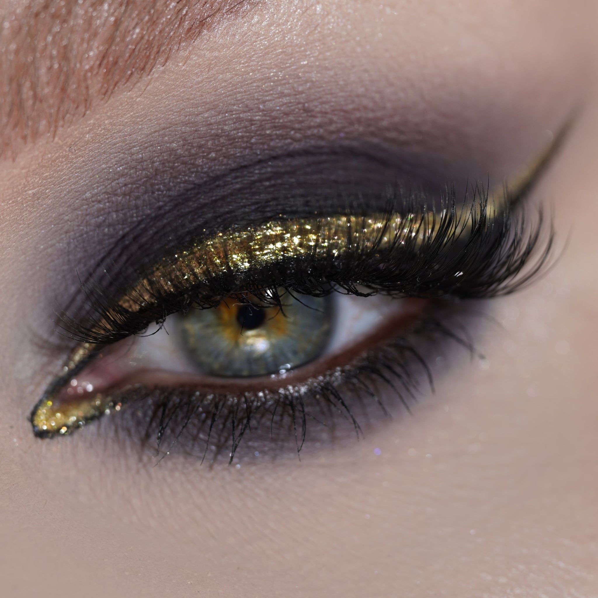 Close-up of an eye with black matte eyeshadow and golden  glitter eyeliner with voluminous lashes.