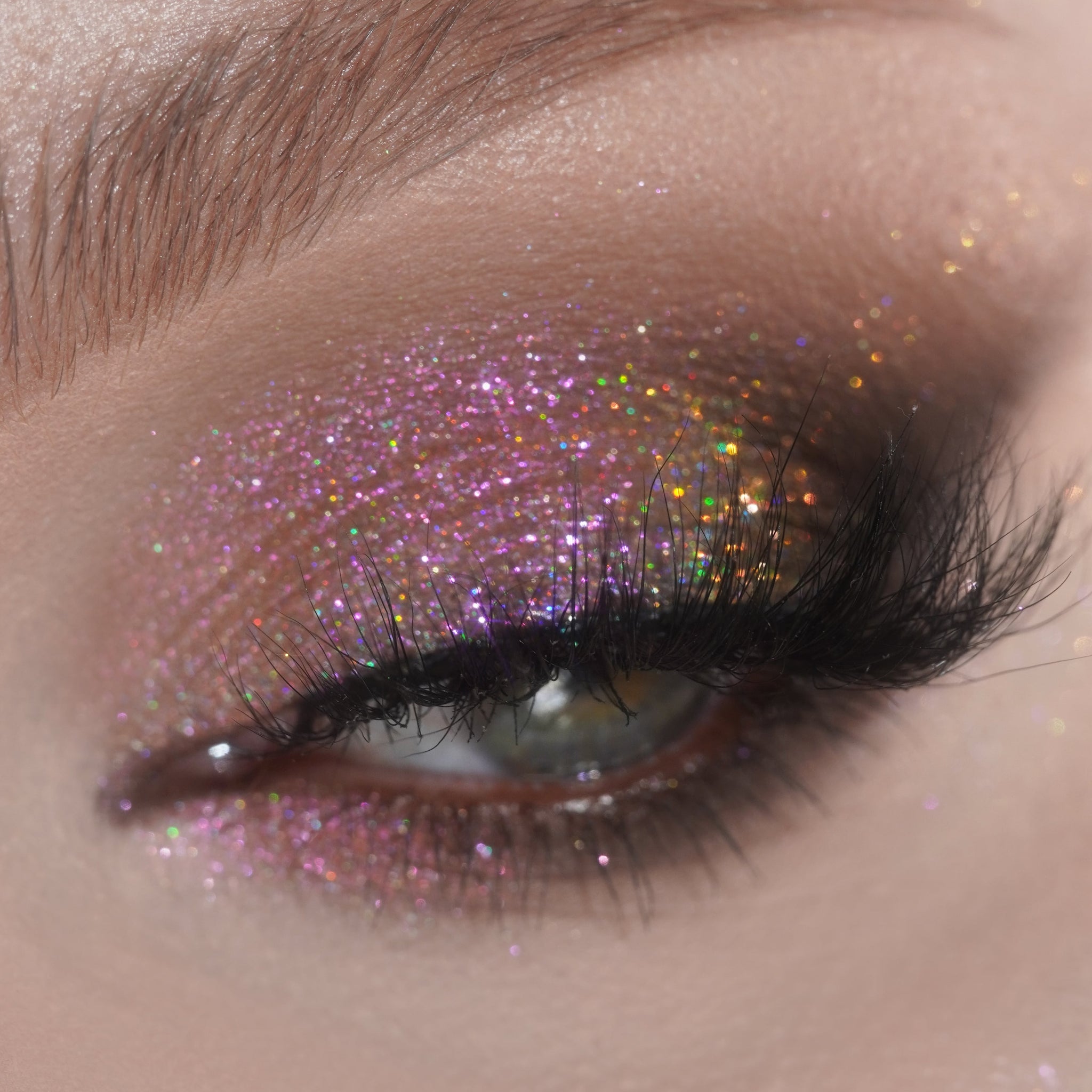 Close-up of an eye with pink holographic glitter eyeshadow, long black lashes, and shimmer on a groomed brow.