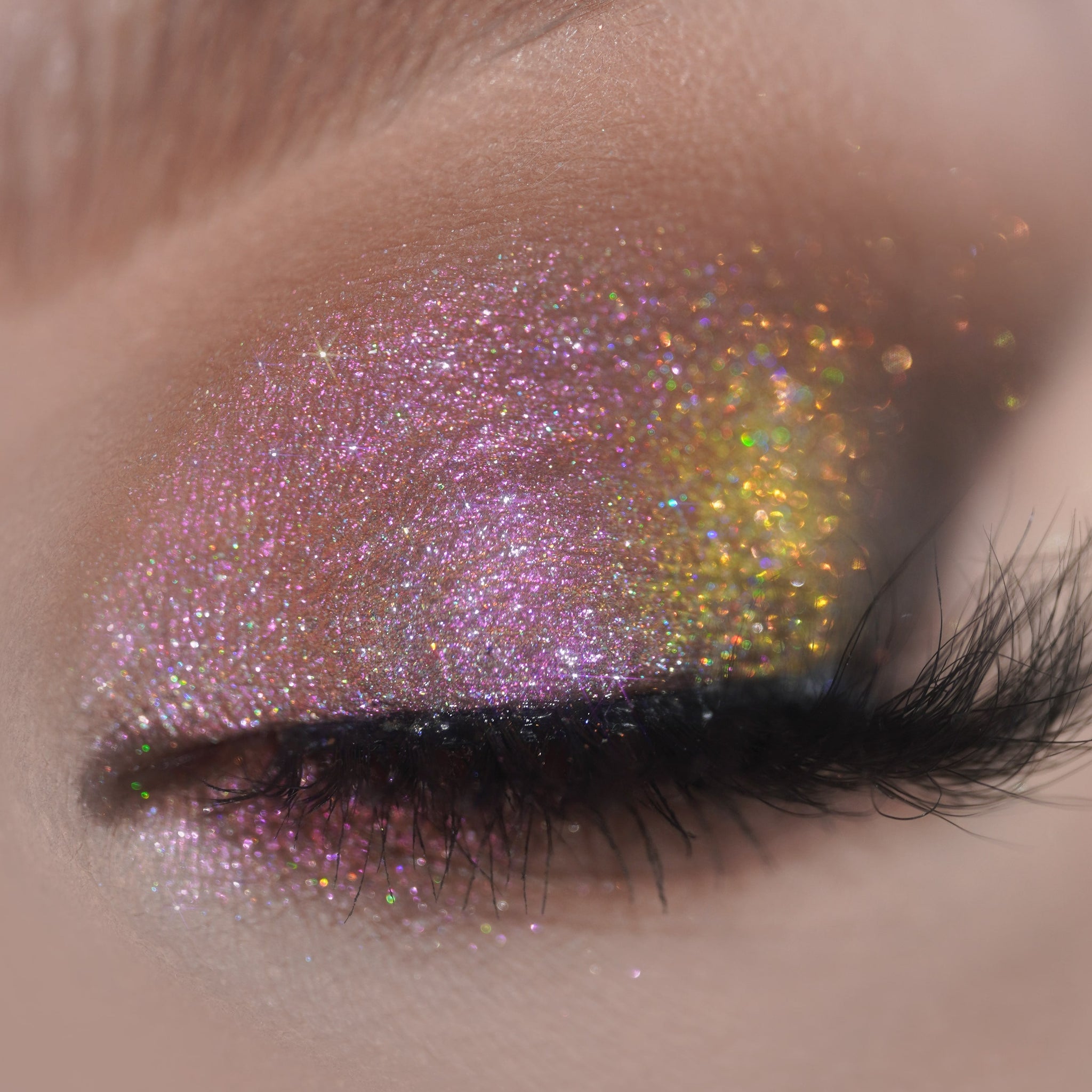 A closed eye shows sparkly pink and gold glitter eyeshadow with long, thick lashes.