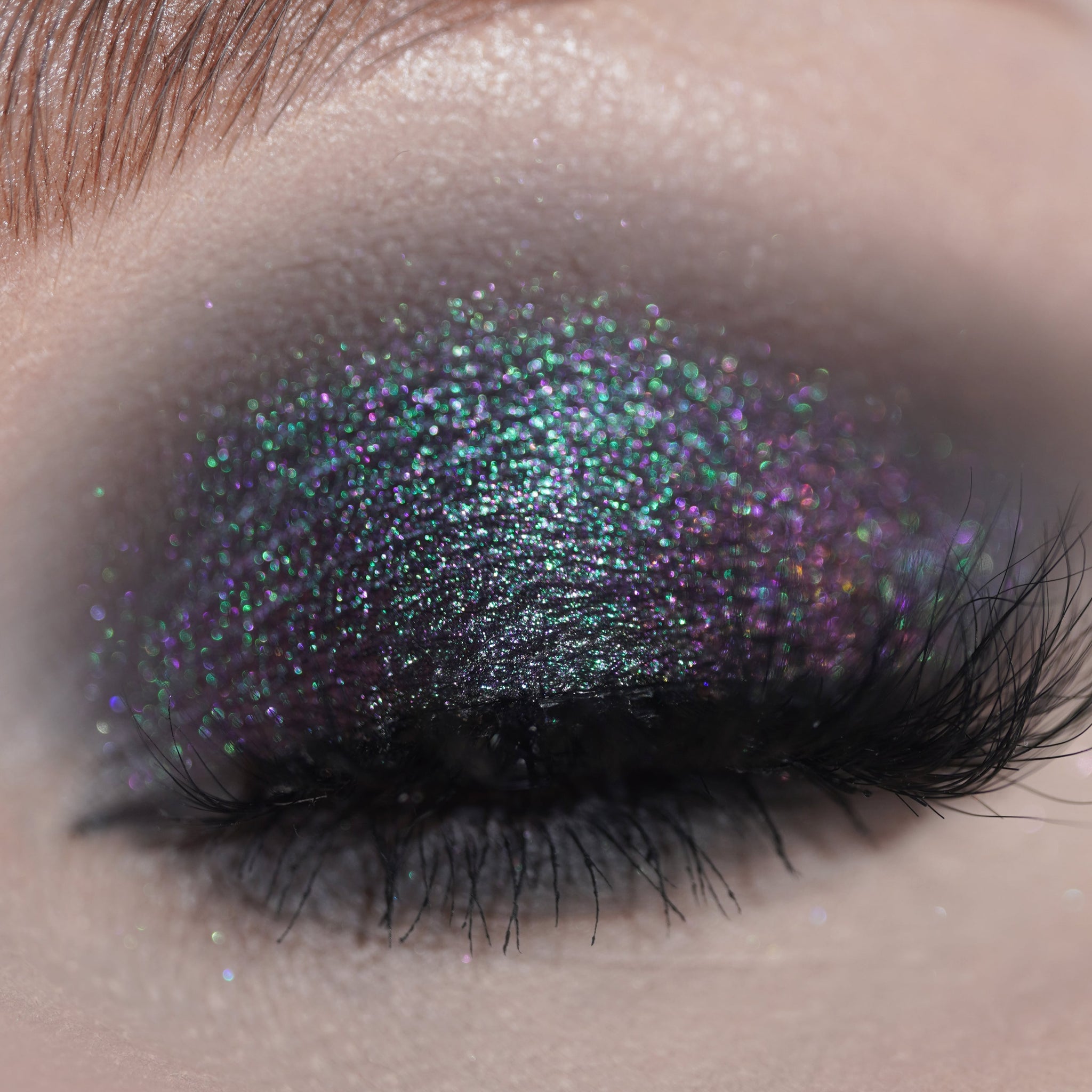 A closed eye shows glittery grey eyeshadow with purple and green specks and long eyelashes.