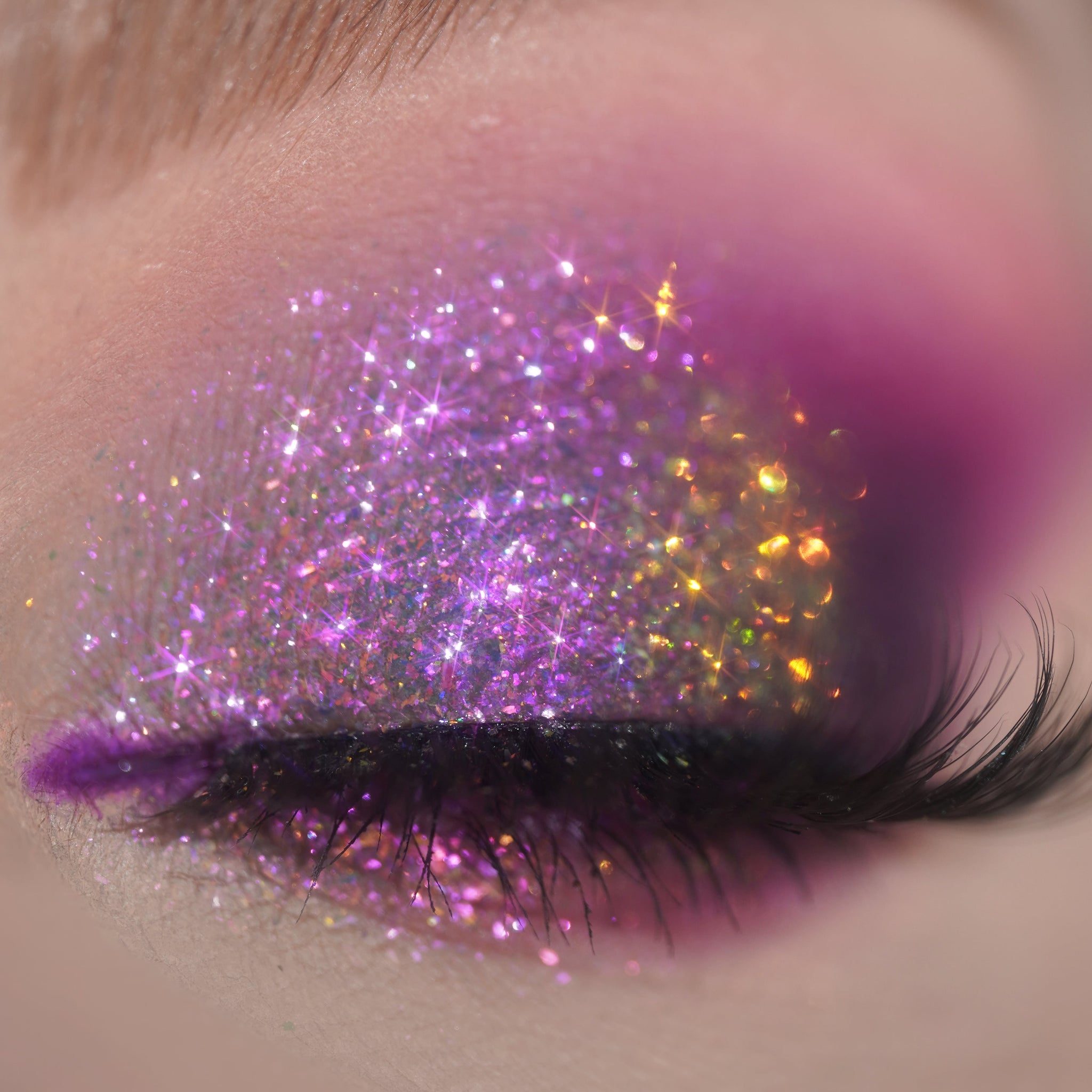 A closed eye with purple and pink glitter eyeshadow, long lashes, and soft pink shimmer on the eyelid.