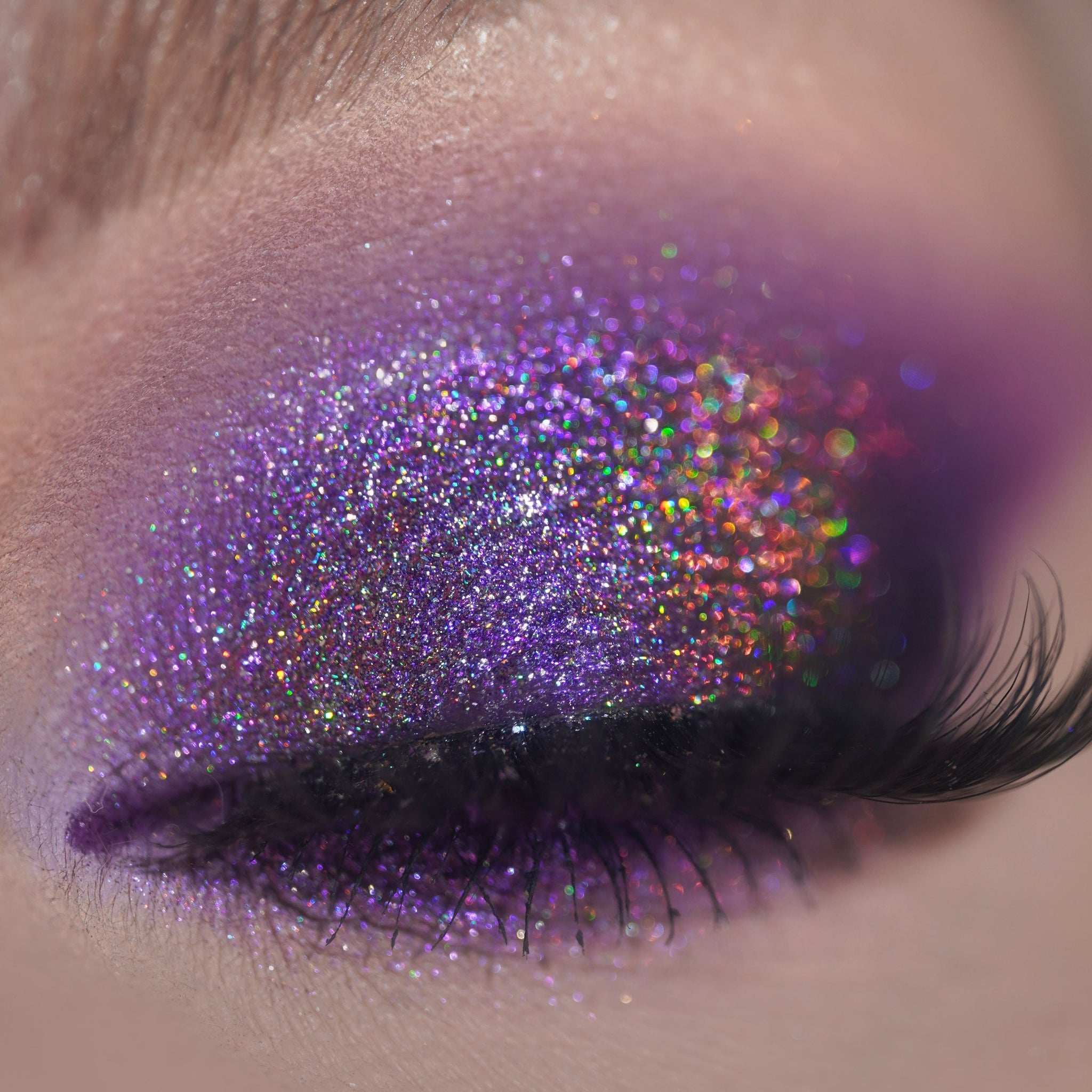 A closed eye shows glittery purple and holographic eyeshadow with long black false lashes.