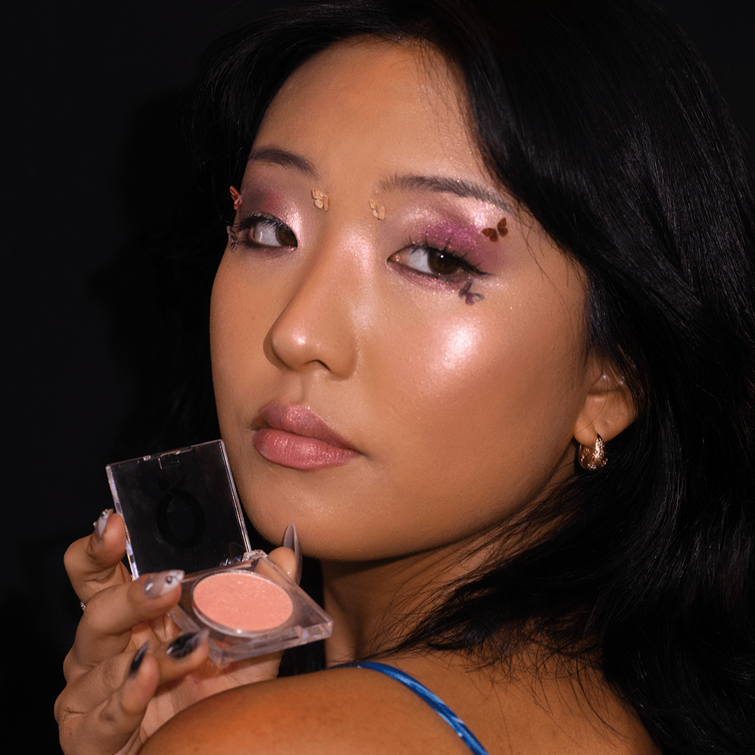 Woman with glossy makeup and floral eyelids holds an open Pressed Pigment Highlighter against a dark background.