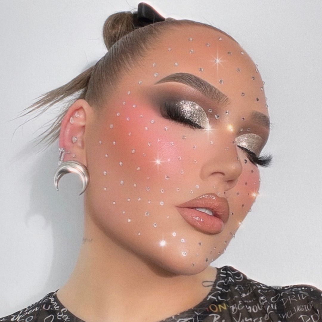 Person with metallic silver eye makeup, nude lips, rhinestones on face, crescent moon earrings, and hair in a sleek bun.