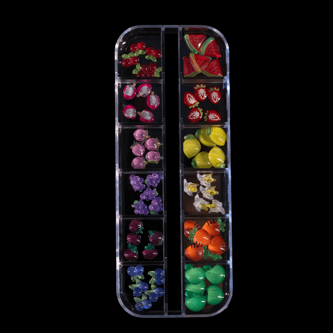 A clear box with separate compartments holds assorted colorful fruit-shaped gems on a black background.