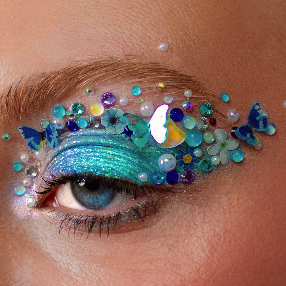 Close-up of an eye with glittery blue eyeshadow, gems, pearls, and butterfly-shaped decorations arranged around it.
