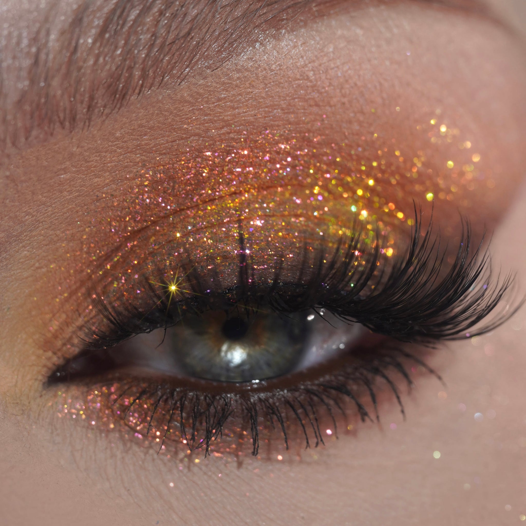 Close-up of an eye with long lashes wearing gold and orange glitter eyeshadow with pink  and yellow accents.