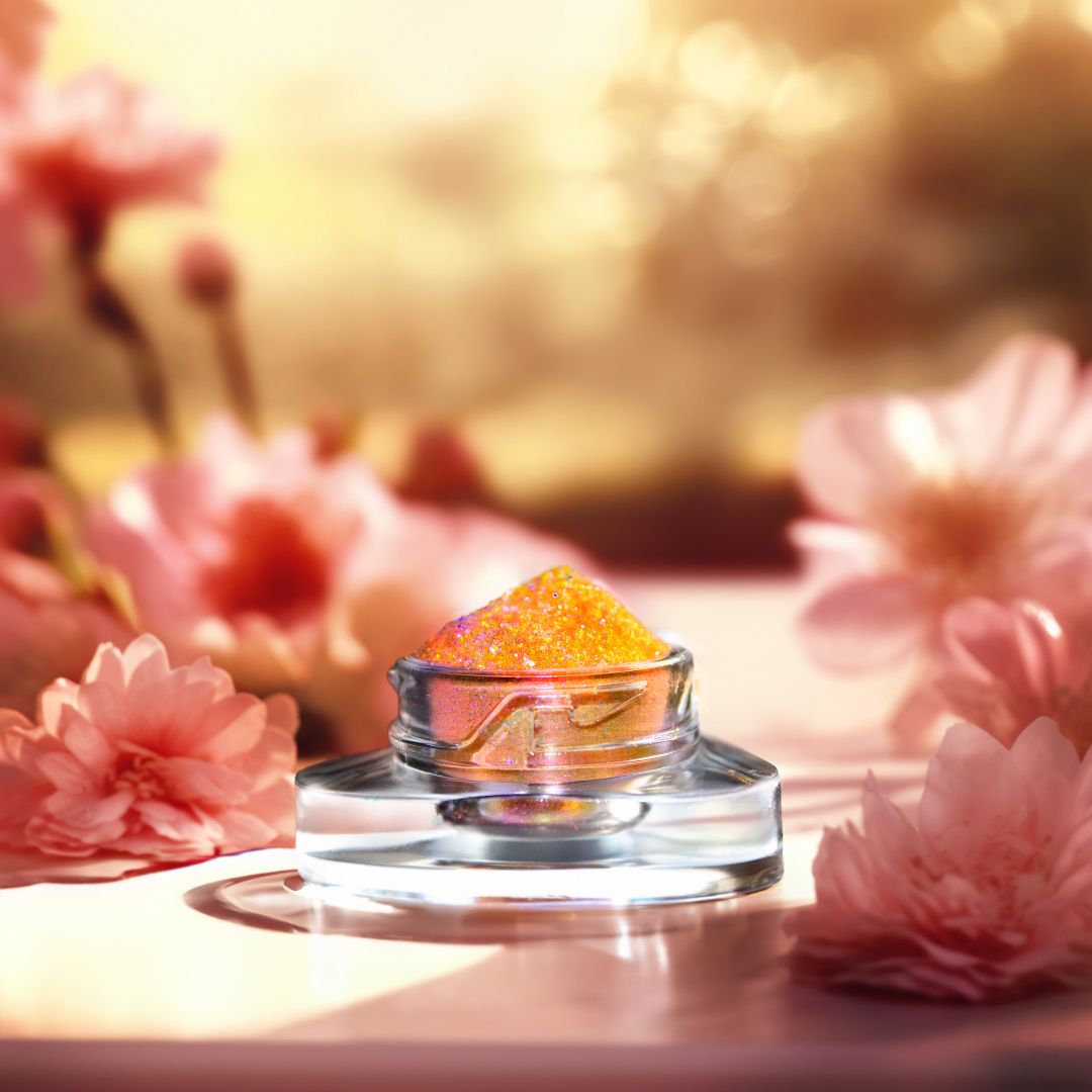 A small jar labeled with orange glitter pigment is placed among several pink flowers with blurry orange background.