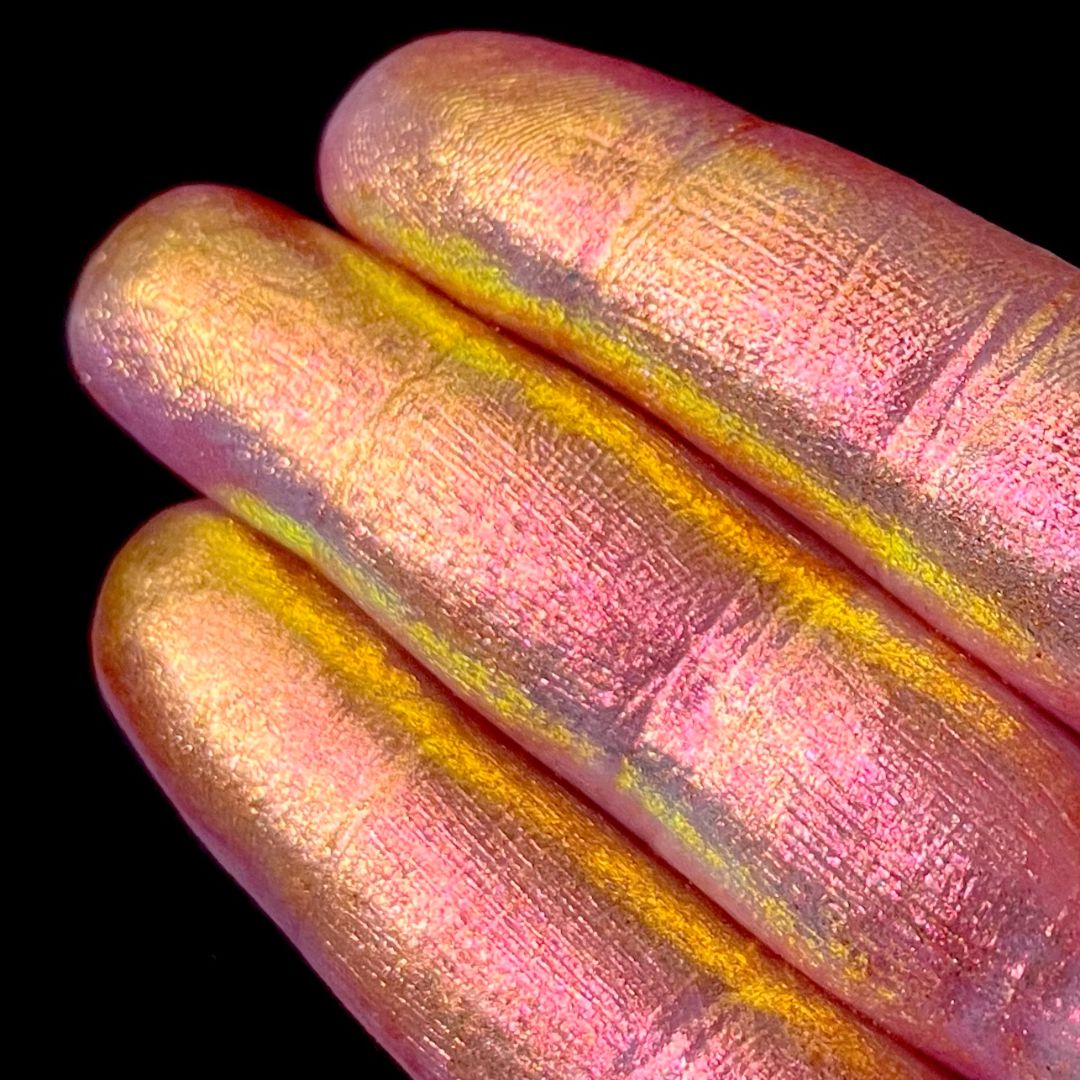 Three fingers display a pink and gold iridescent shimmer pigment on their nails polish against a black background.