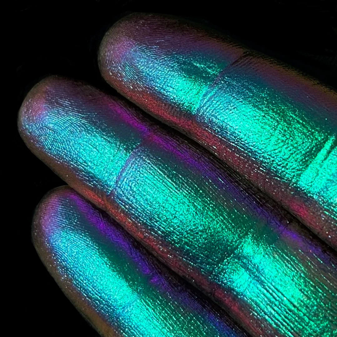 Close-up of three fingers with shimmering green and blue pigment on their tips against a black background.