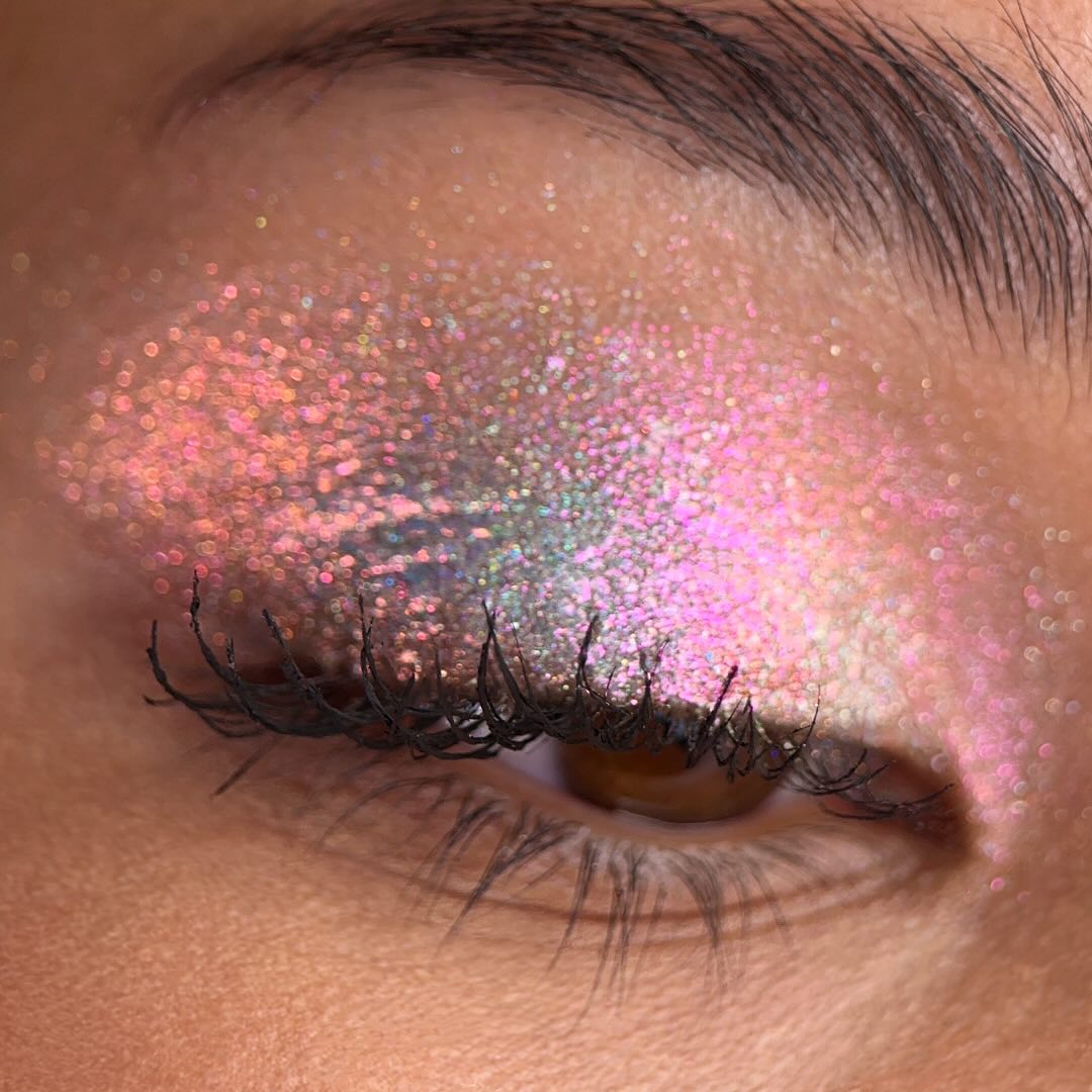 A closed eye with long lashes is shown with iridescent pink glitter eyeshadow on the eyelid and surrounding skin.
