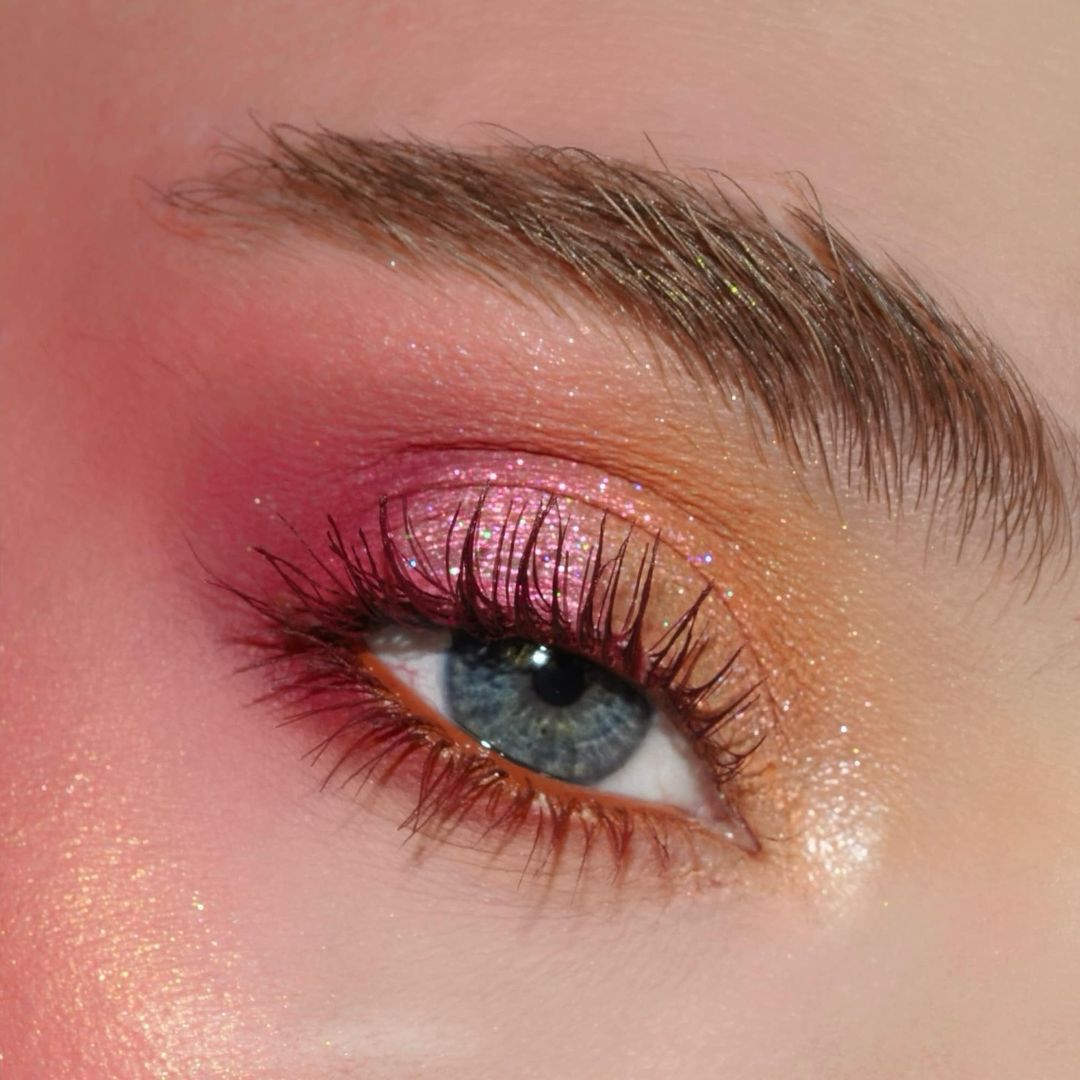 Close-up of a blue eye with pink and gold shimmery eyelids, long dark lashes, and a neatly shaped eyebrow.