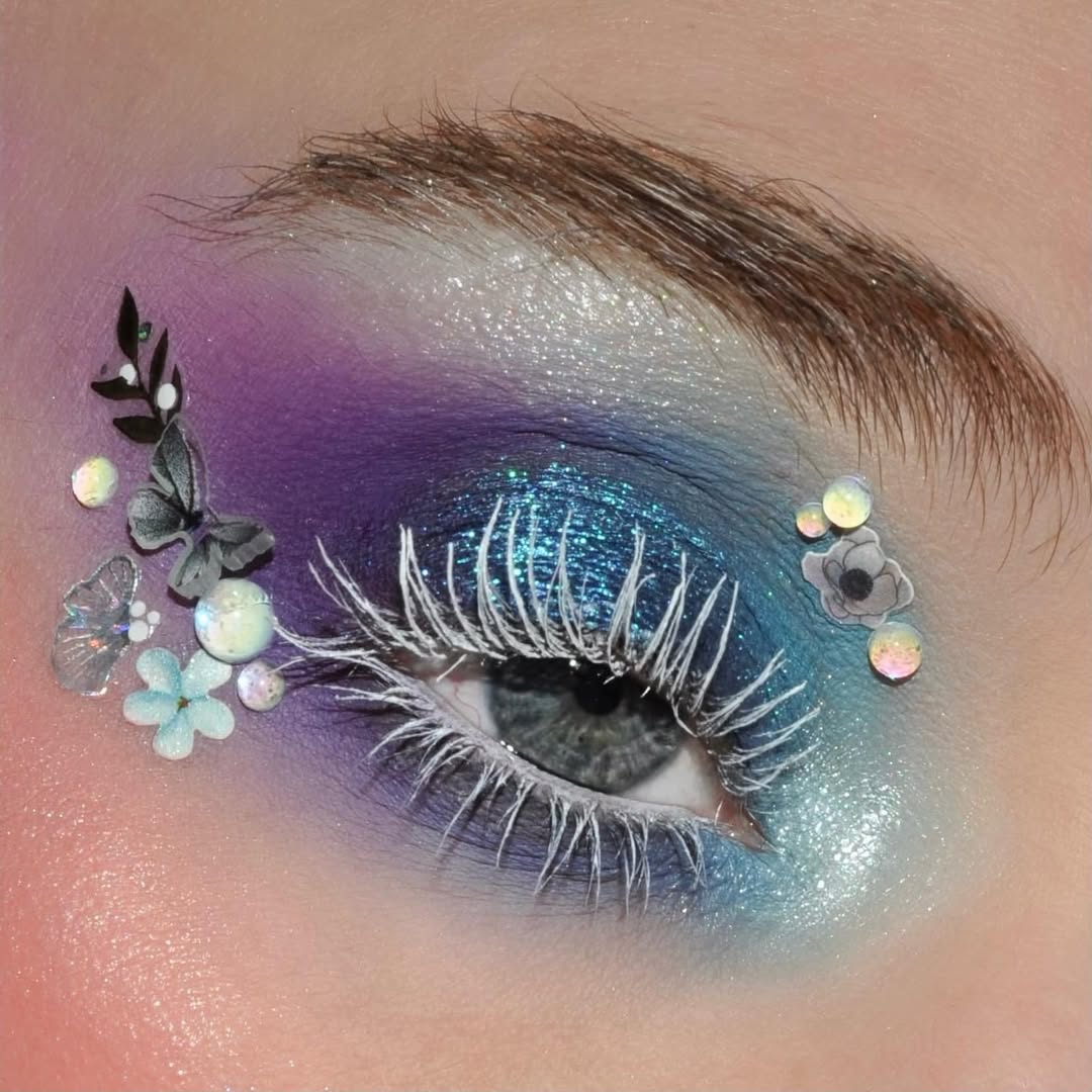 Close-up of an eye with blue and purple sparkly eyeshadow, small floral gems, and white Arctic Ice mascara.